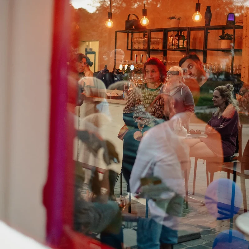 Door het raam gezien: een volle zaal en de toog van BarOH in warm licht